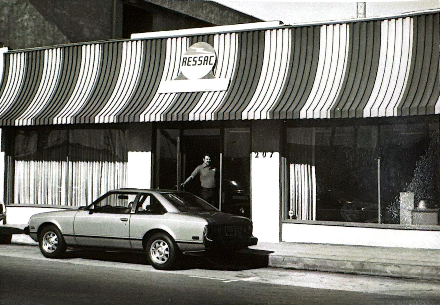 A storefront with the name RESSAC (old name), in the background a man entering, and an old car parked on the street, in an urban setting at night.