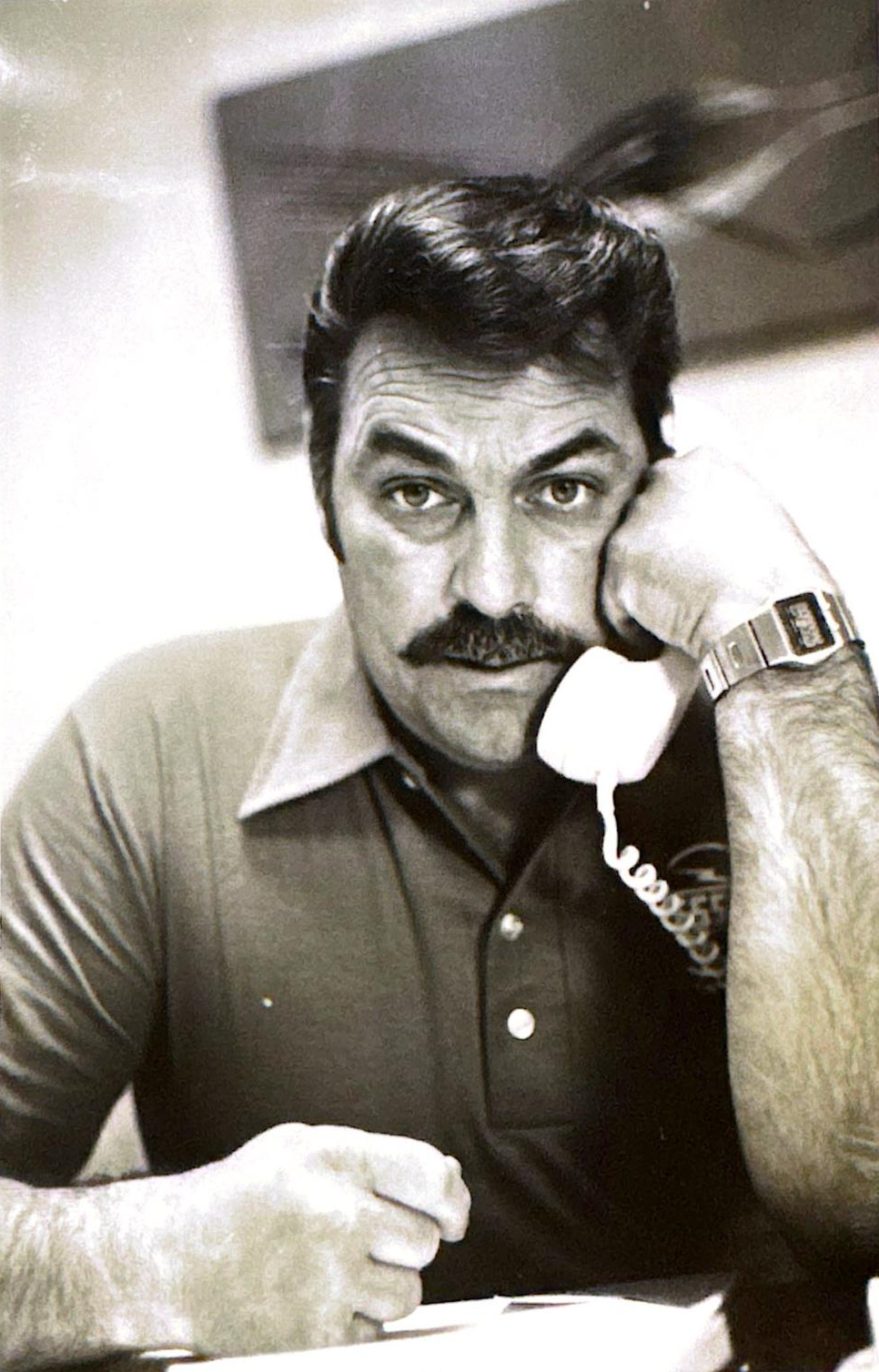 Man with mustache, dark hair and thoughtful expression, holding old telephone and wearing polo shirt, indoors, in black and white.